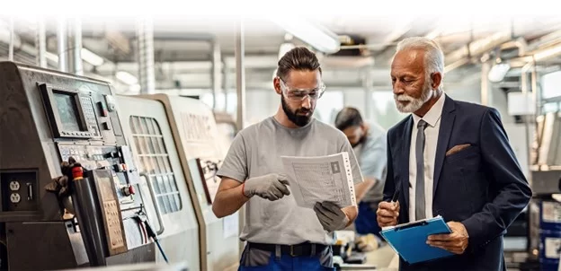 A manufacturing engineer and a manager reviewing a CNC machine programming sheet in a well-lit factory, with a CNC machine visible in the background.